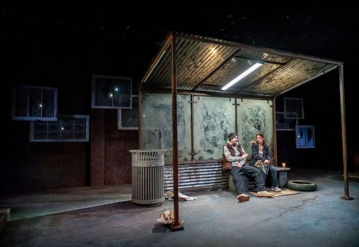 Under a metal corrugated shelter, a man and a woman sit talking.
