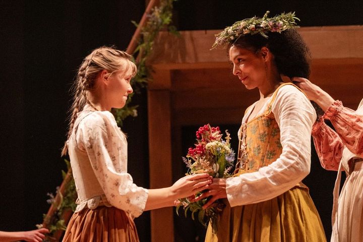 A young girl hands a bouquet to a woman who wears a floral crown and is getting ready for her wedding. They smile at each other.