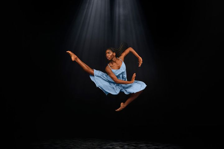 A ballerina wearing a blue dress leaps in the air with one leg extended, high off the ground against a black backdrop.