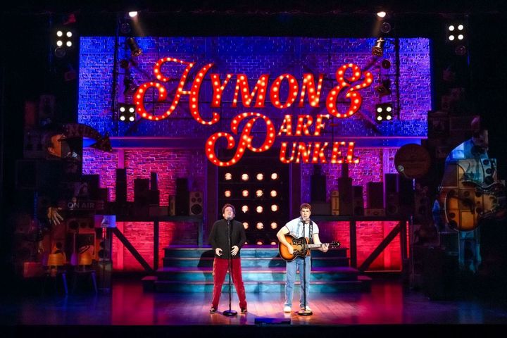 Two men stand on a stage singing while one plays the guitar.