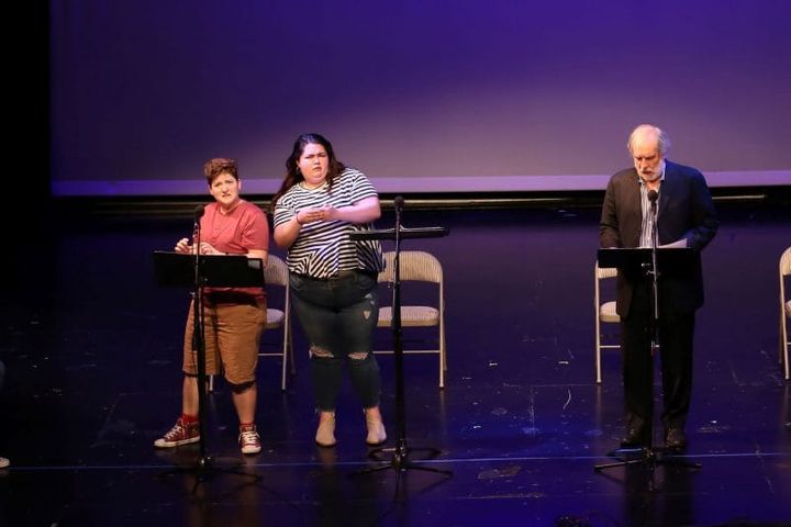 Three people stand on stage with microphones and music stands in front of them doing a play reading.