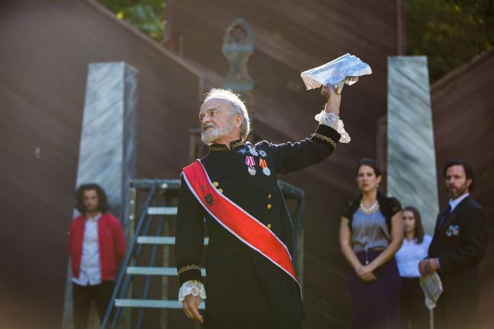 A man wearing a dress military uniform speaks to a crowd looking angry.