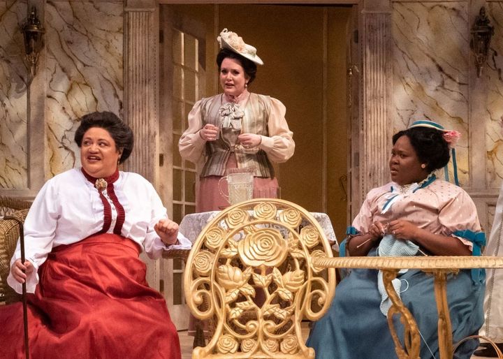 Two women sit wearing late 1880s outfits they look incredulously at someone. A third woman stand behind them.