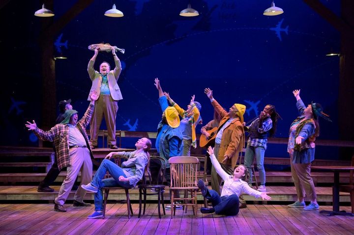 A group of men and women all have their arms open wide as they sing.  A man in the back holds up a fish above his head.