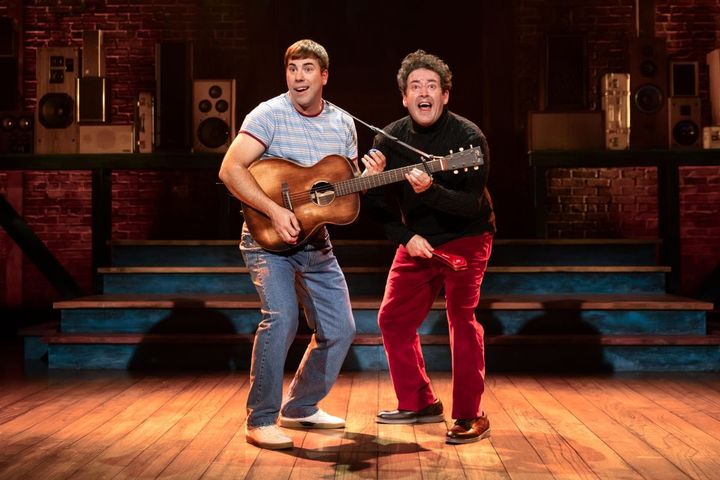 Two men stand on a stage singing. One plays the acoustic guitar.