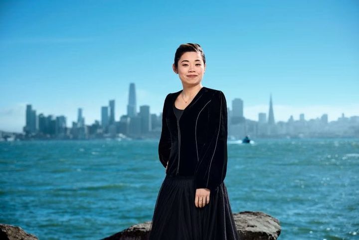 Elim Chan stands on rocks in front of the bay with water and the city skyline of San Francisco behind her.