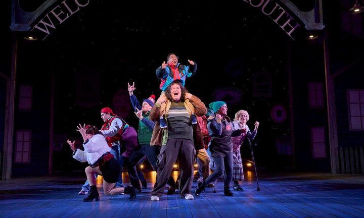 A large group of people are in a group, the ones in the back are dancing expressively. Two boys are in the front, one on the other's shoulders.