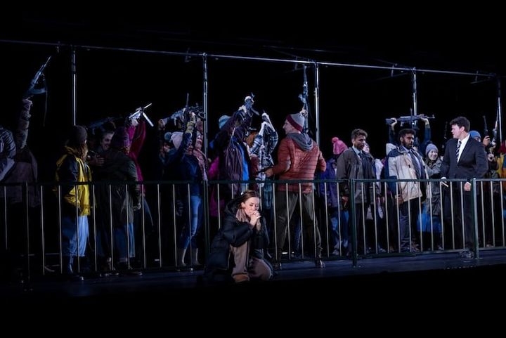 A woman kneels praying in front of a barricade behind which people shout.