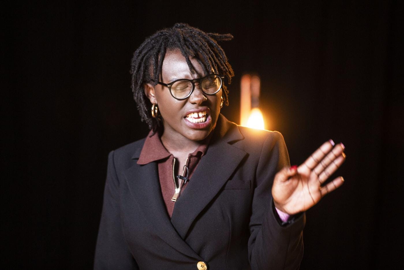 A black person with short locs, wearing a black blazer, black glasses and brown top midway through a performance.