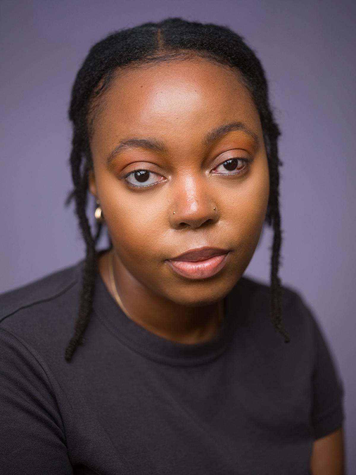 A South African person with brown eyes looking into the camera, she is wearing a black top with gold jewellery. 