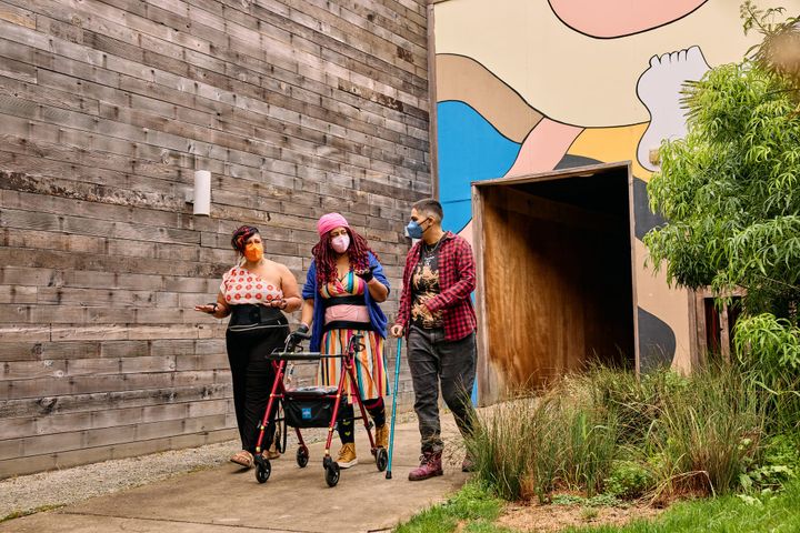 Three masked disabled people of color chat while strolling through an outdoor courtyard.