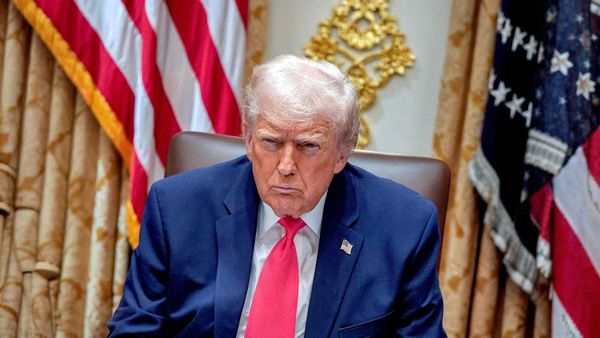 Donald Trump seated at a desk in a formal setting with U.S. flags behind him.