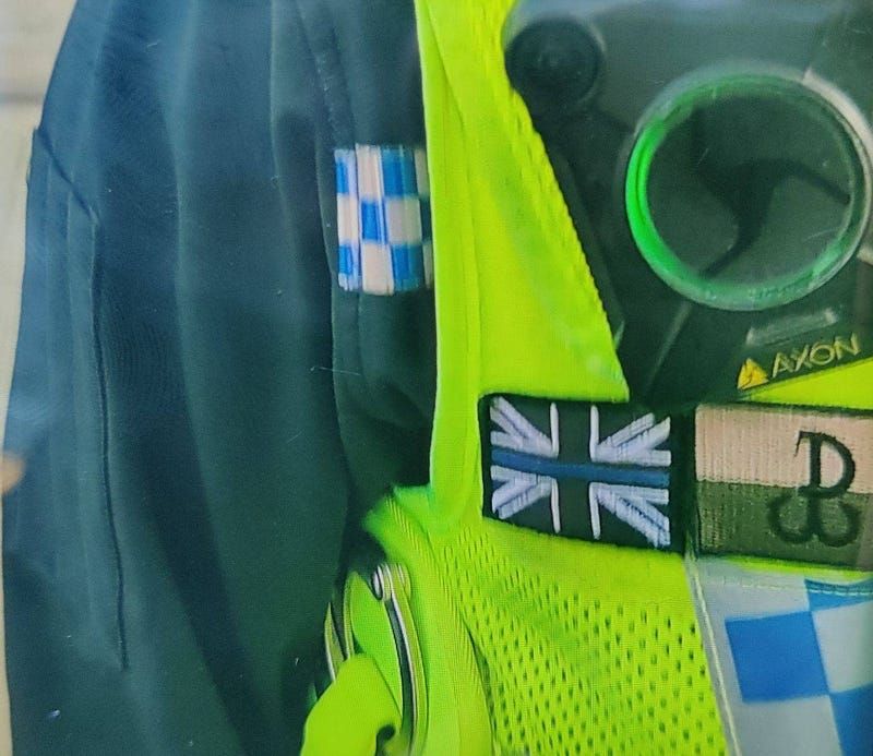 Police officer close up yellow police vest, thin blue line over a union flag patch and a tan and olive horizontally striped p