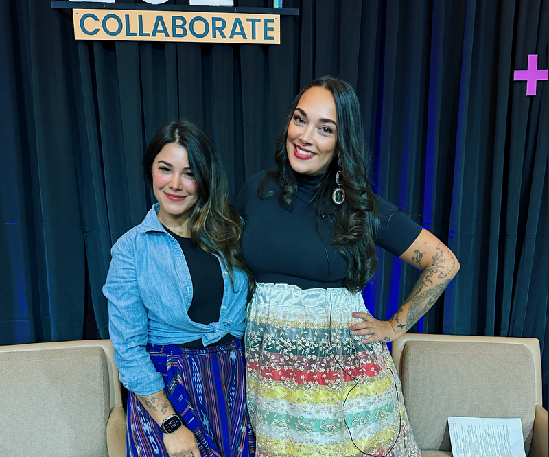 Latin woman on left with colorful native print blue skirt, chambray shirt and black undershirt. Standing with another taller