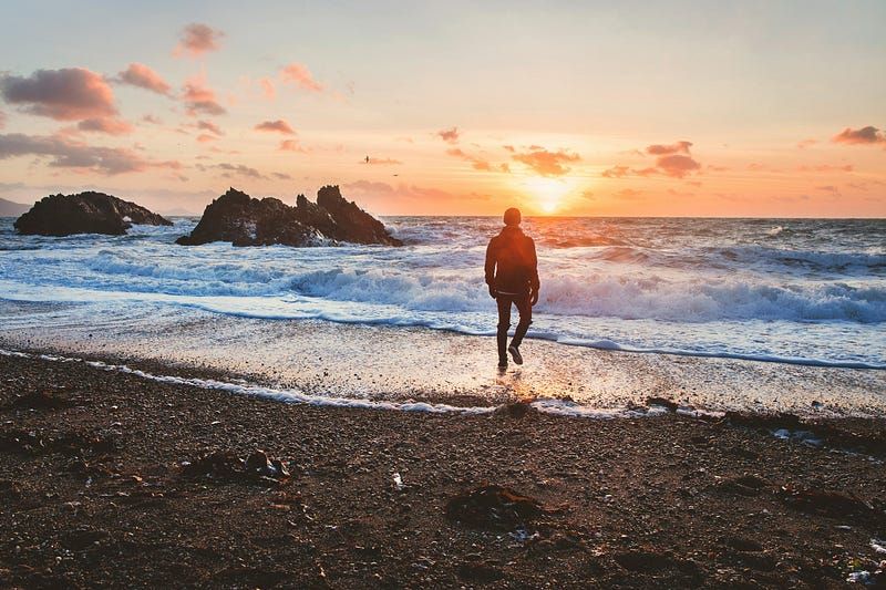 A person is running along a pebble beach towards the sea at sunset. The sun, low in the sky, casts a warm glow across the sce