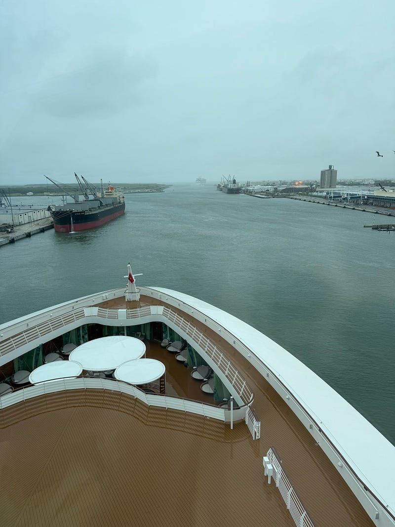 A view from the top deck of a cruise ship docked at a port on a rainy, overcast day. The deck appears wet from the rain, and