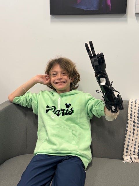 Photo of child wearing upper limb prosethetic. They are sitting on a couch giving the peace sign with the prosethetic and smi