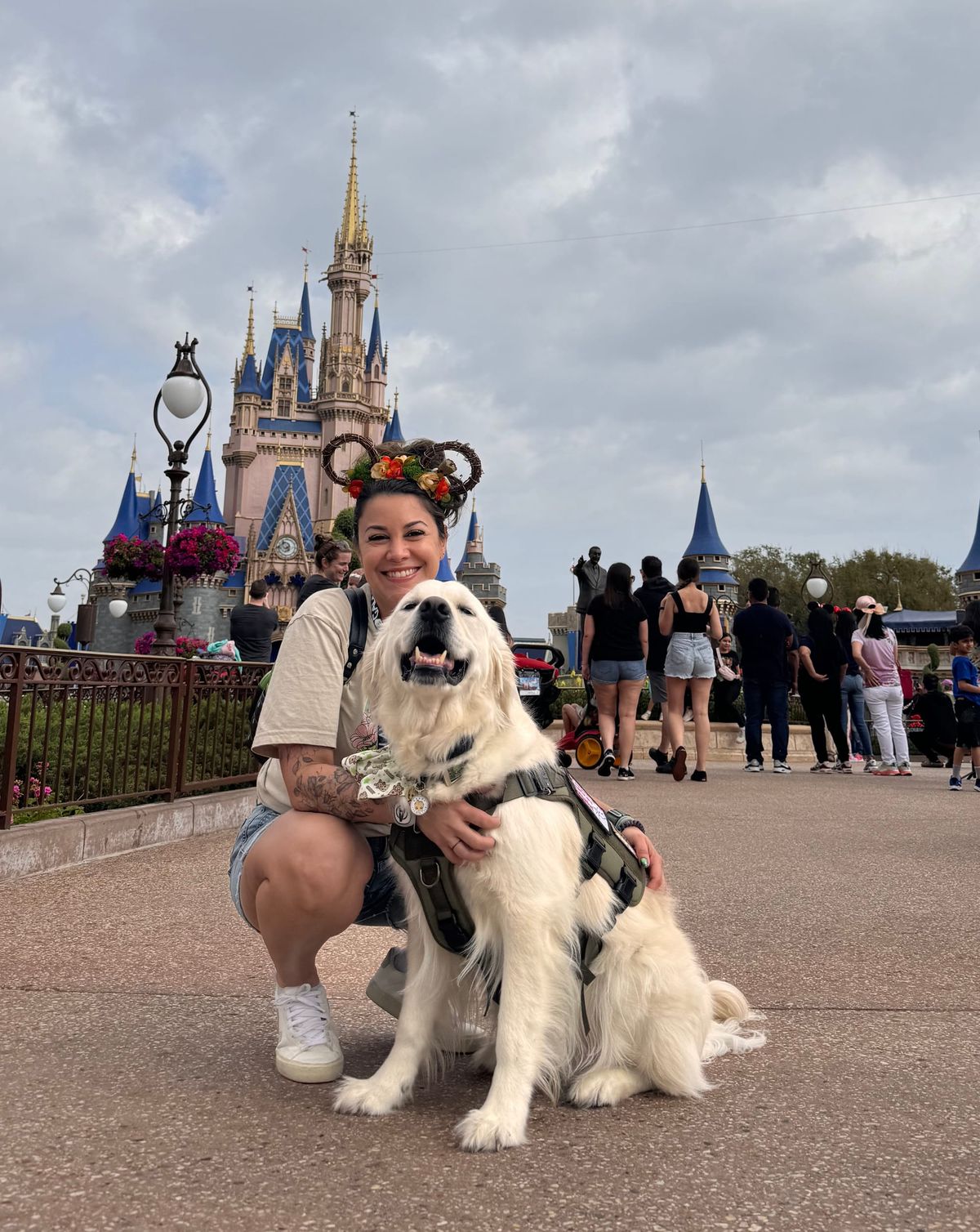 Woman with flower Minnie ears kneels beside her service dog in front of Cinderella’s Castle at Disney World. The dog wears a harness and looks happy.