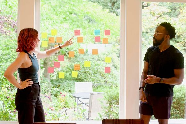 Two colleagues engaged in a brainstorming session, standing by a large window covered in colorful sticky notes. One person is