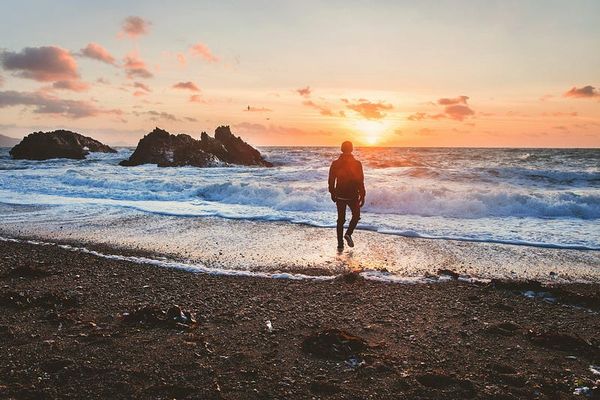 A person is running along a pebble beach towards the sea at sunset. The sun, low in the sky, casts a warm glow across the sce