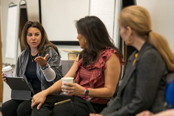 One woman on the left is gesturing while speaking, and the other two women are listening attentively. They all hold coffee cu