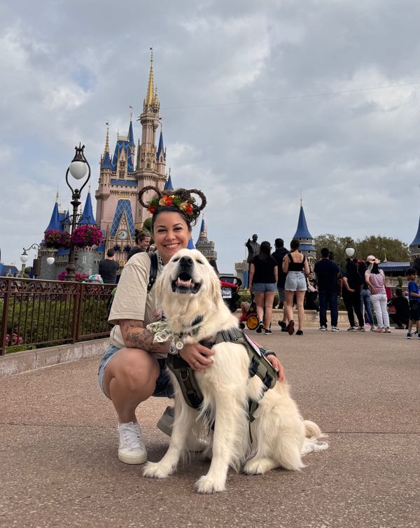 Woman with flower Minnie ears kneels beside her service dog in front of Cinderella’s Castle at Disney World. The dog wears a harness and looks happy.