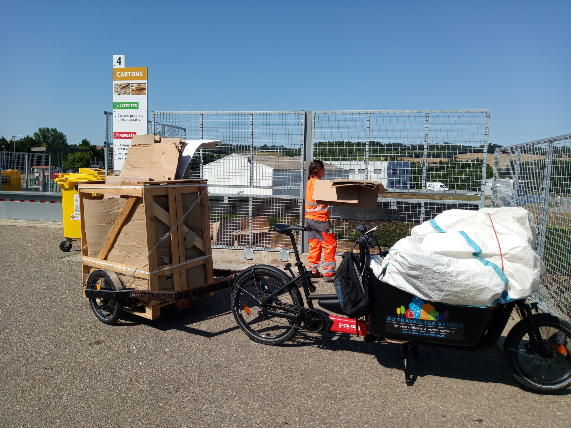 Un vélo cargo Douze G4, tranportant un sac de polystyrène expansé dans sa caisse et tirant une remorque Bicylift a conduit une caisse palette transportant des cartons à recycler dans une déchèterie.