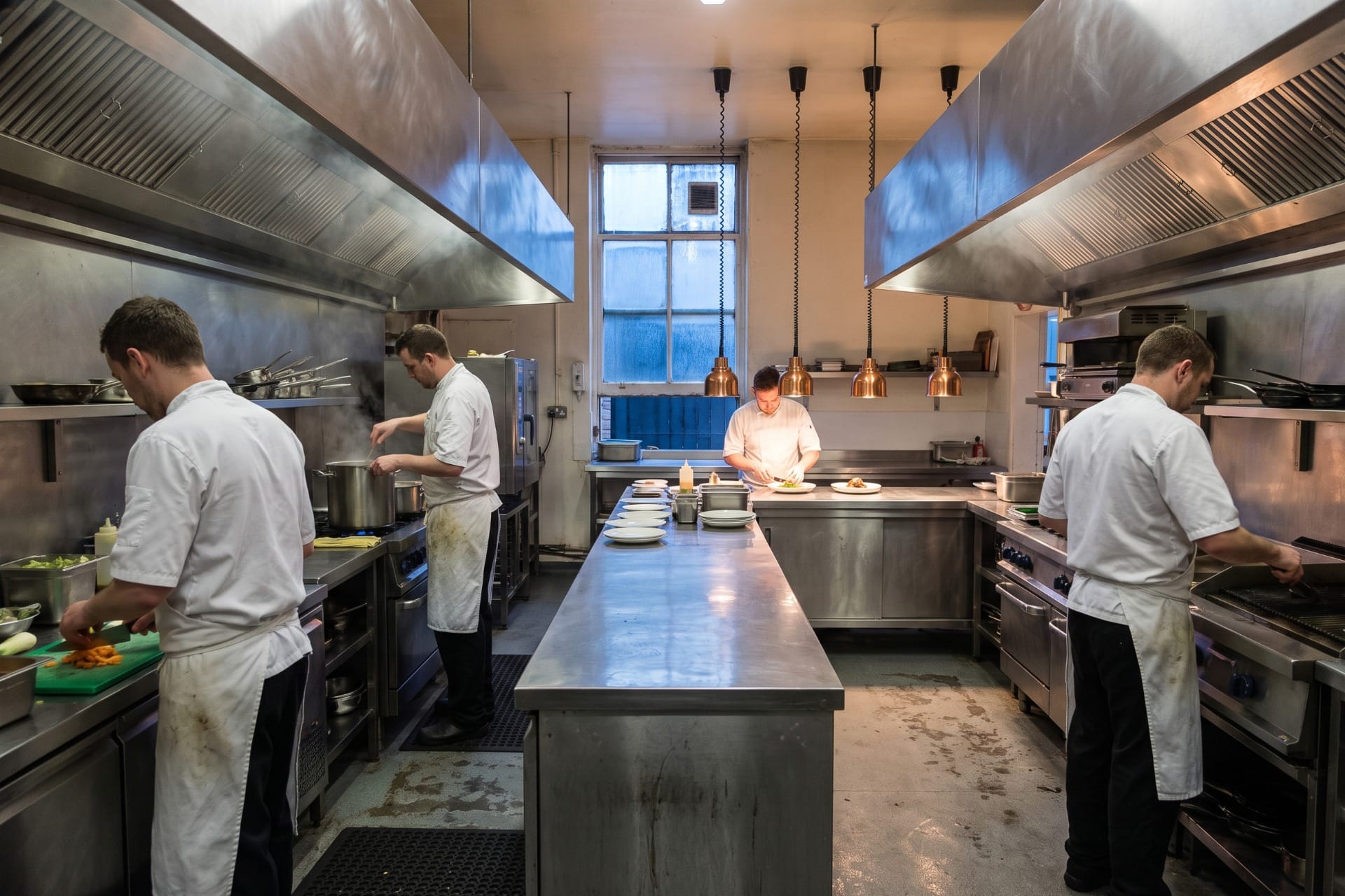 A long commercial kitchen with chefs working at stations spread far apart under two extraction canopies