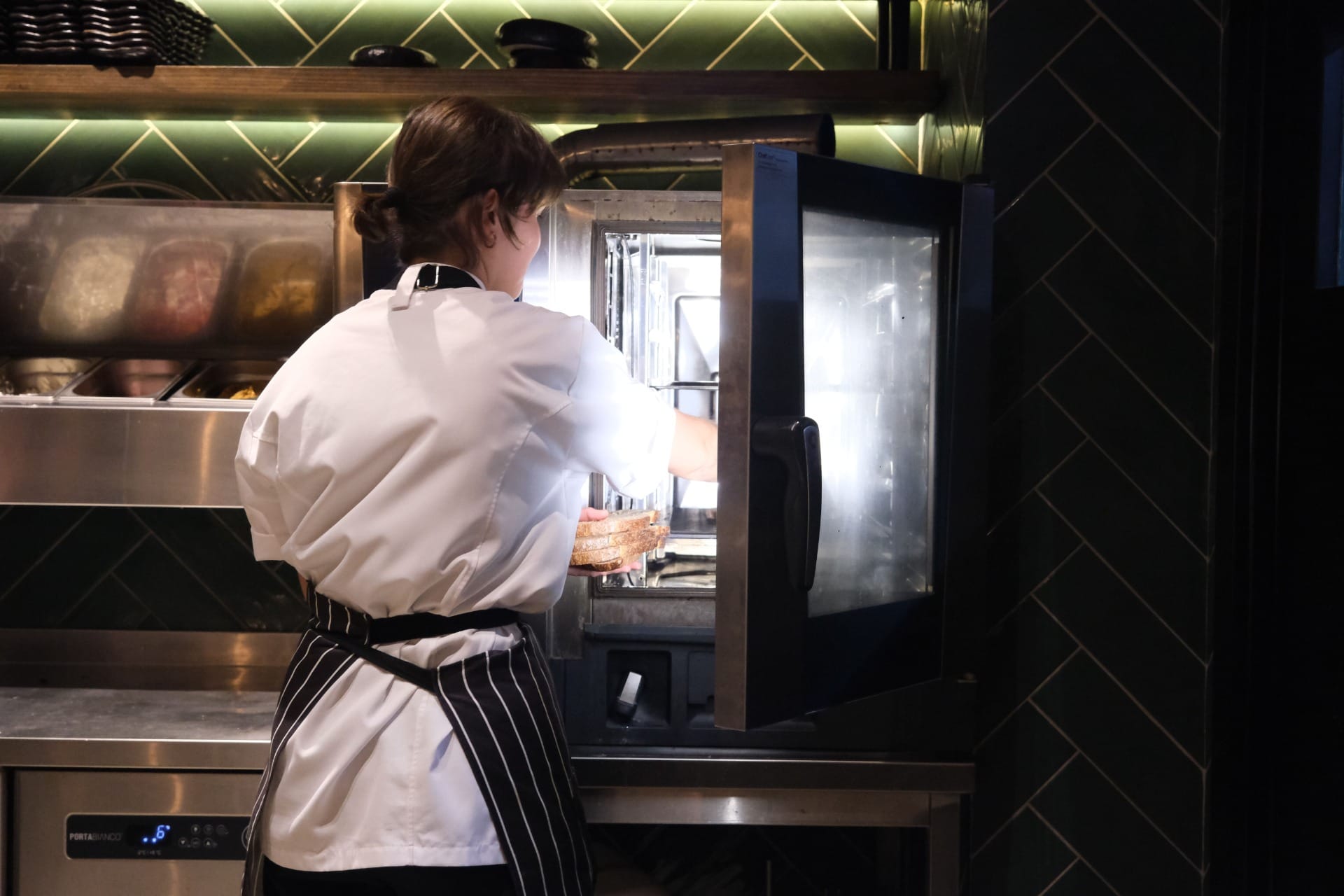 A chef loading trays into a combination oven during service in a tiled commercial kitchen
