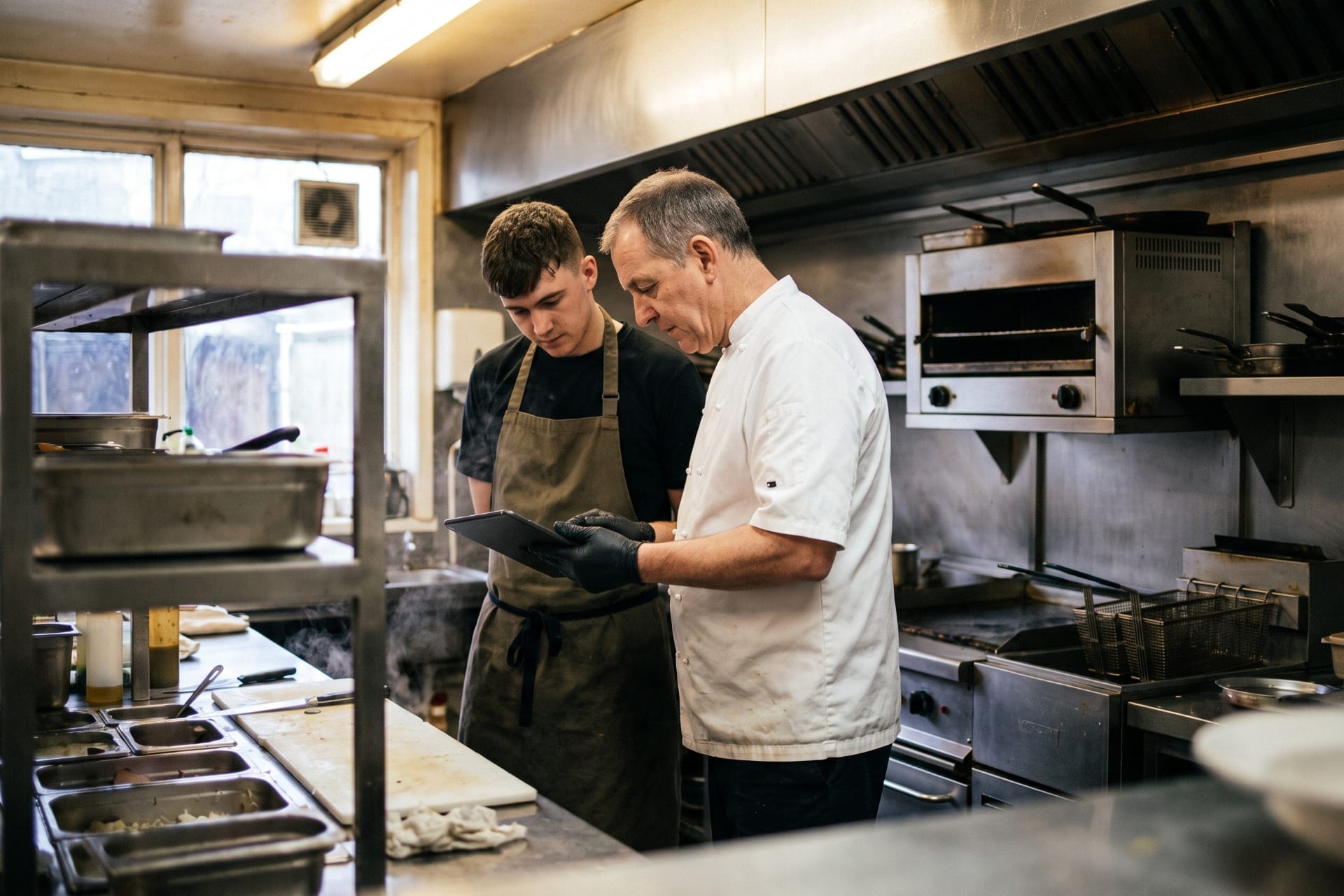 A head chef showing a younger cook something on a tablet at the bench during service in a working commercial kitchen