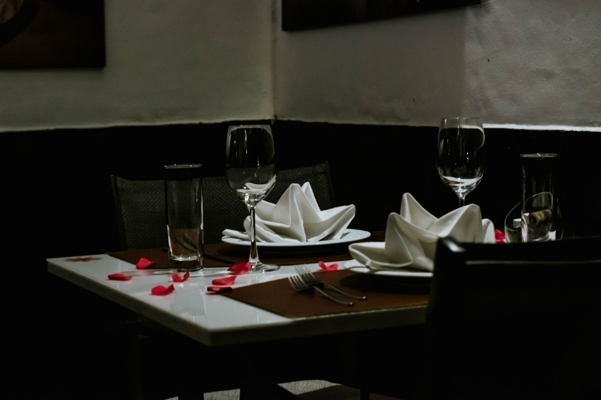 A restaurant table set for two with folded napkins, wine glasses and scattered rose petals