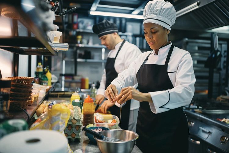 Two chefs working side by side at a prep bench in a commercial kitchen with a mixing bowl between them.