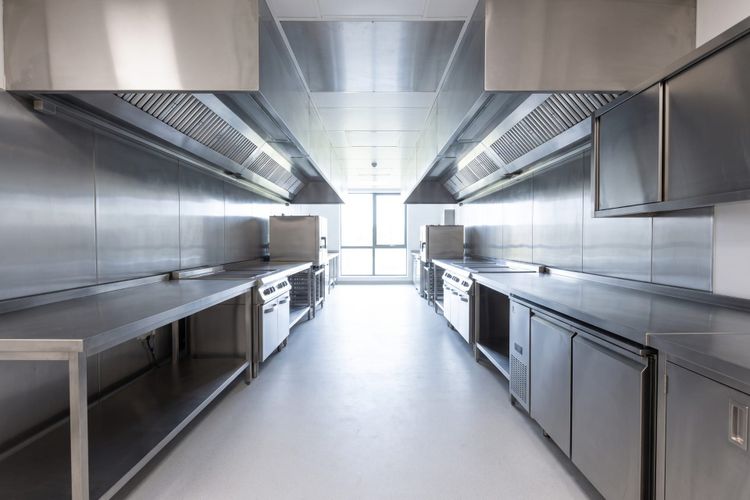 Modern commercial kitchen photographed down its length, with a clear central walkway, stainless steel benches along both sides and extraction canopies running overhead.