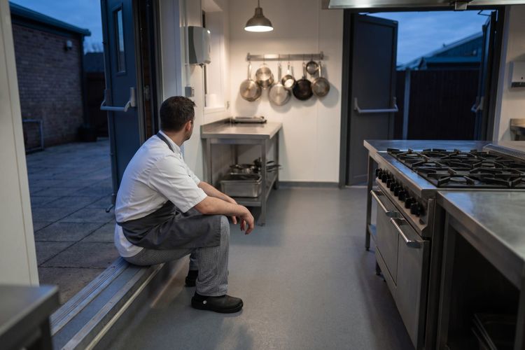 A chef sitting alone on the step of an open kitchen back door at dusk, the empty kitchen behind him.