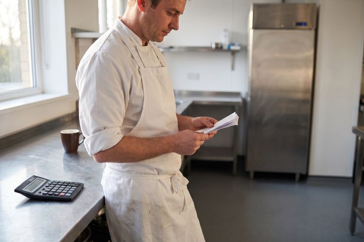 A chef reading a stack of papers at a stainless kitchen bench, a calculator and a mug on the bench beside him.
