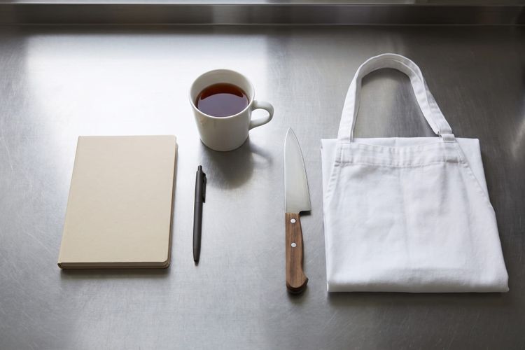 A notebook, pen, mug of tea, chef knife and folded apron laid out on a stainless steel kitchen bench, seen from above