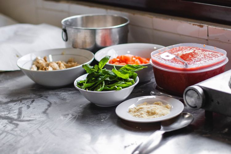 Bowls of prepped ingredients laid out on a stainless kitchen bench, with green leaves, chopped tomato and grated cheese.