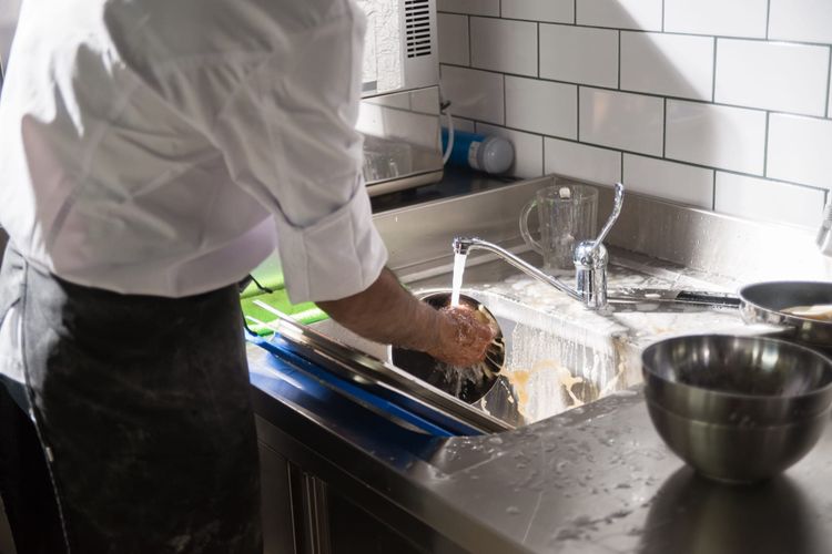 A chef in whites washing a pan under running water at a stainless commercial sink with white tiled walls.
