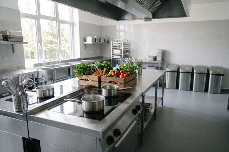 Pans working on flat induction hobs in a daylit stainless kitchen, waste bins lined up along the wall behind.