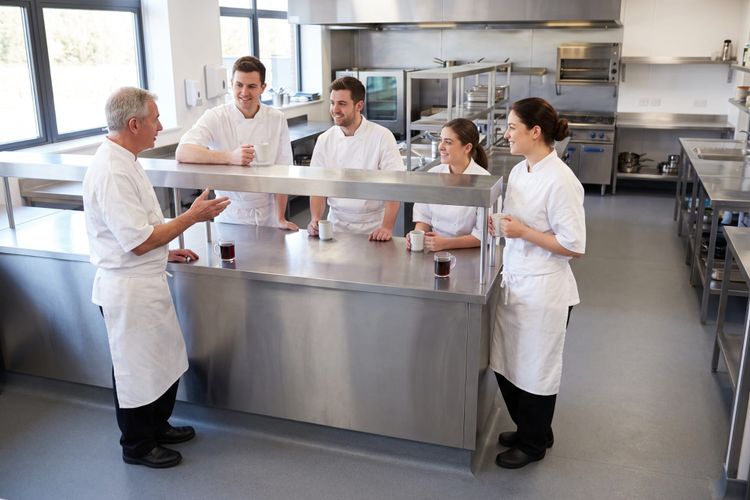 Five kitchen staff in whites gathered round a stainless pass with mugs, one talking and the rest listening.