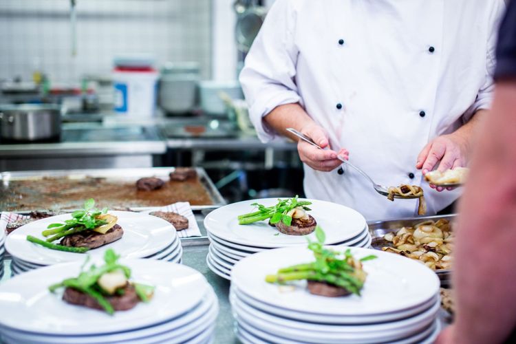Plated fillet steaks lined up on the pass with stacks of warm plates as a chef finishes the garnish