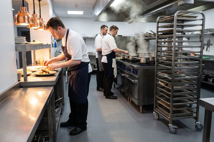 A chef plating a dish at the pass under copper heat lamps while two others work the range behind.