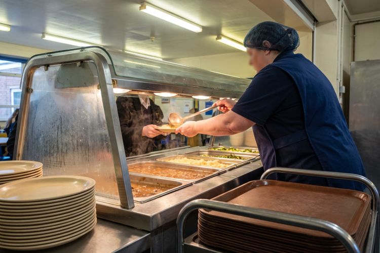 A school kitchen assistant serving mashed potato and gravy from the hot counter onto a plate at a school servery