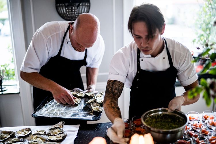 Two chefs in aprons working side by side over a tray of oysters and a bowl of green dressing.