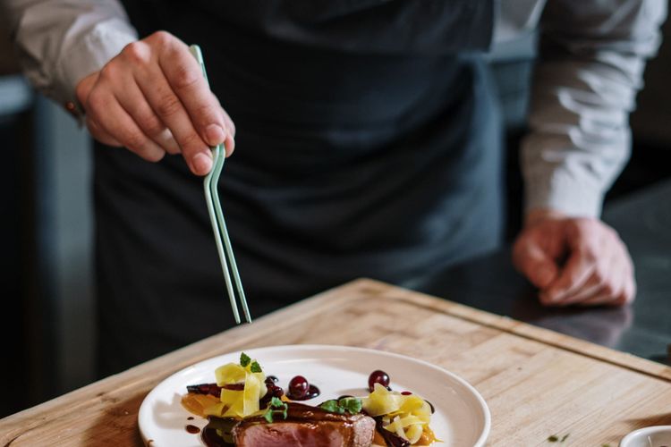 A chef in an apron finishing a plated dish with tweezers on a wooden board.