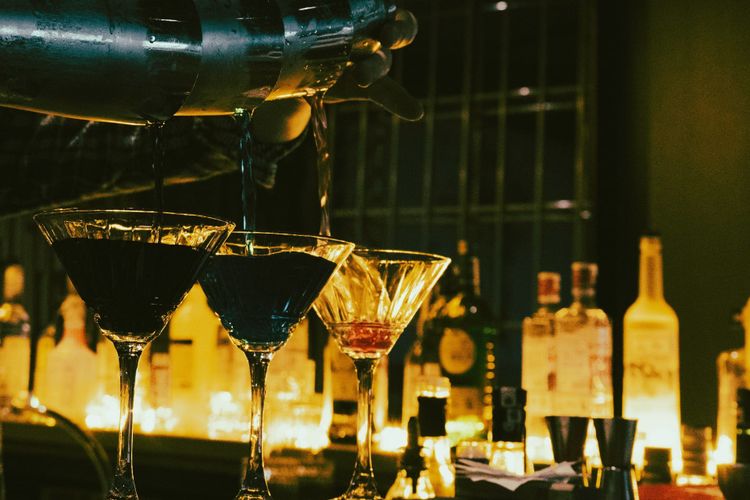 A bartender pouring a cocktail from a shaker across a row of coupe glasses at a warmly lit back bar