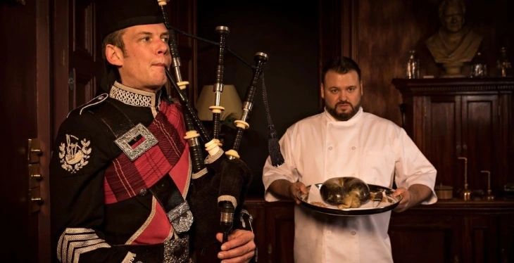Piper in full highland dress playing the bagpipes beside a chef in whites carrying a haggis on a platter.