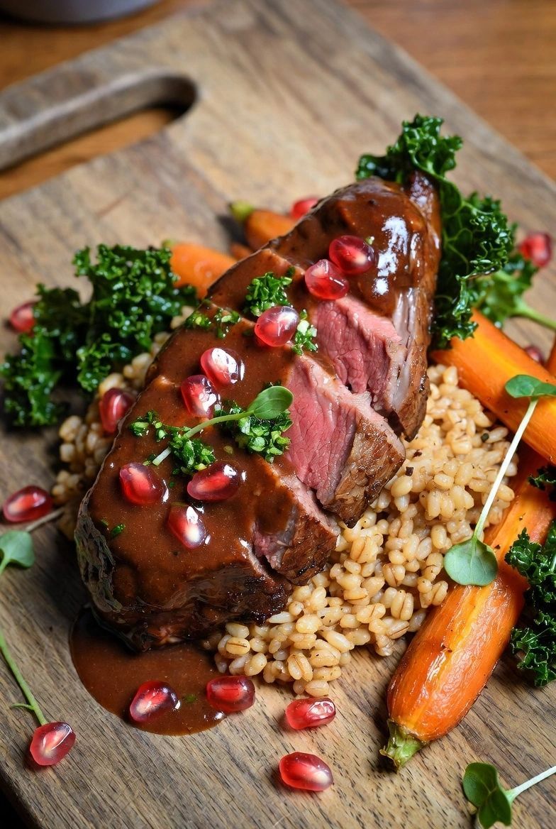 Sliced miso and honey glazed lamb rump on a bed of grains with carrots and berries on a dark wooden board.