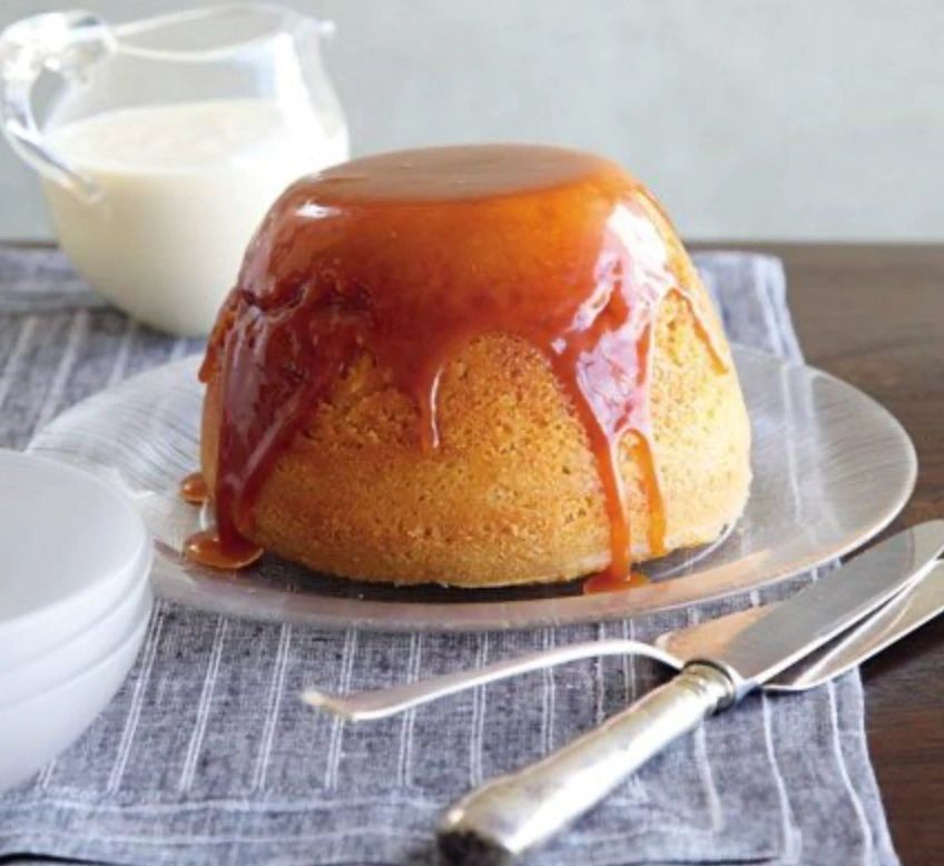 Steamed sponge pudding with butterscotch sauce running down the sides, a jug of cream and spoons alongside.