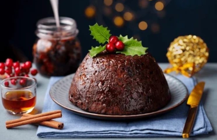 Christmas pudding topped with holly on a plate, with a jar of brandy butter, a glass and cinnamon sticks alongside.